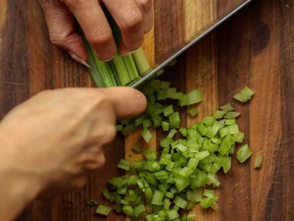 A close-up of hands chopping fresh celery stalks into small pieces on a wooden cutting board with a large knife.